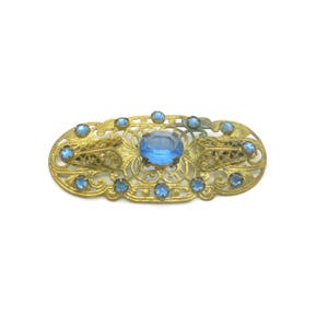 May include: A gold-toned filigree brooch with a light blue oval-shaped rhinestone in the center. The brooch is decorated with smaller light blue rhinestones around the edge.