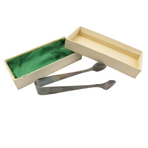 Könnte beinhalten: Eine silberfarbene Vintage-Zuckerzange mit muschelförmigen Enden, neben einer offenen rechteckigen Schachtel. Die Schachtel ist beige mit einem grünen Satinfutter und goldenem Rand.