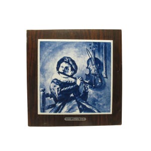 May include: A blue and white Delft tile with a framed image of a young person playing a flute. The tile is framed in dark wood and has the text "1620 - J. STEEN - 1679" at the bottom.