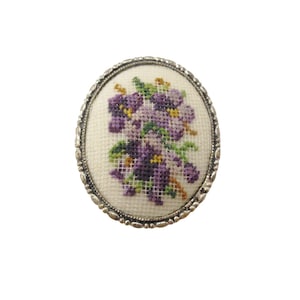 Puede incluir: Broche ovalado con un diseño floral bordado a punto de cruz. El bordado muestra un ramo de flores moradas con hojas verdes y detalles amarillos sobre un fondo crema. El broche está enmarcado por un borde plateado ornamentado.