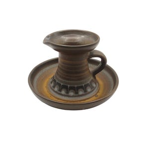 May include: A brown ceramic pitcher with a handle and spout, set on a matching saucer. The pitcher has a textured, ribbed design with a decorative pattern around the base. The saucer has a yellow and brown gradient.