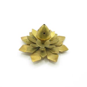 Può includere: Un bruciatore di incenso in metallo dorato a forma di fiore di loto con una parte superiore conica. Il bruciatore ha più strati di petali e un piccolo foro al centro della parte superiore.