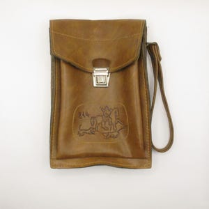May include: A brown leather satchel with a flap closure and a silver-colored clasp. The bag features a hand strap and an embossed design of a person on a horse. The bag has a rectangular shape and is made of a textured leather material.