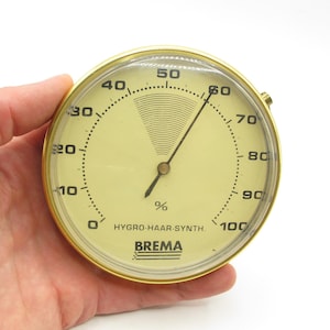 May include: A circular hygrometer with a gold-tone metal frame. The cream-coloured face features black numerals and a needle showing the humidity percentage. The text "HYGRO-HAAR-SYNTH." and the brand "BREMA" are printed on the face.