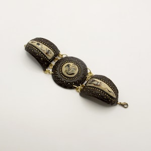 May include: A brown bracelet with three decorative sections. The sections are connected by gold-coloured chains. Each section has a dark brown base with a textured pattern and cream-coloured accents. The centre section is circular, while the other two are curved.
