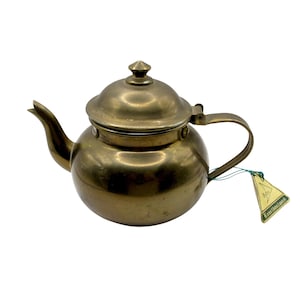 May include: A vintage brass teapot with a lid and handle. The teapot has a curved spout and a small tag attached with green string. The tag has text on it.