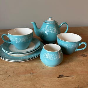 May include: A teal tea set with white floral patterns. The set comprises a teapot, two teacups, saucers, and a sugar bowl. The teacups and saucers are stacked on a wooden surface. The teapot has a lid and a handle. The sugar bowl is round.