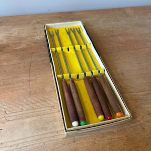 May include: A vintage fondue fork set in a cardboard box. The set includes six forks with wooden handles and stainless steel tines. The handles are capped with white, green, yellow, red, and orange tips. The box has a yellow interior.