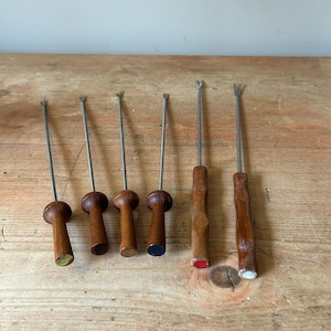 May include: A set of six vintage fondue forks. Each fork has a wooden handle and metal tines. The handles are brown, with some having painted tips in green, blue, red, and white. The forks are displayed on a light-coloured wooden surface.