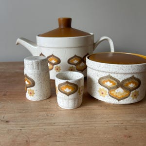 May include: A vintage ceramic tea set comprising a teapot, sugar bowl, salt shaker, and cup. The set has a speckled white finish with brown and yellow floral patterns. The teapot and sugar bowl have brown lids.