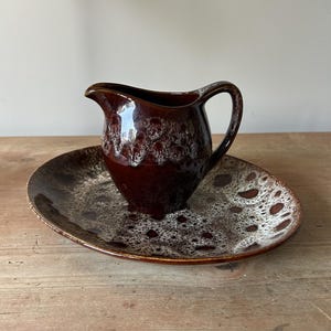 May include: A vintage ceramic jug and plate set. The jug is a deep brown with a white speckled pattern and a curved handle. The oval plate has a similar speckled design in brown and white. The set is displayed on a wooden surface.