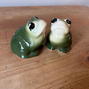 May include: Two ceramic frog ornaments. The frogs are green with white bellies and black eyes. They are positioned on a wooden surface. These are likely decorative items.