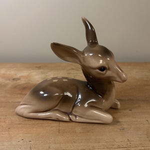 May include: A ceramic fawn figurine in a resting position. The fawn is brown and tan with white spots. The figurine has a glossy finish and is placed on a wooden surface. The figurine is a decorative item.