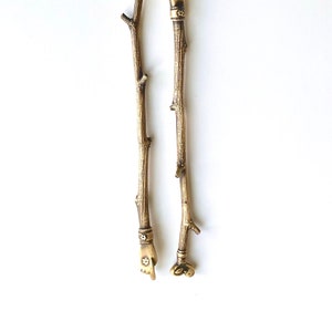 Może przedstawiać: Dwa złote metalowe patyczki z ozdobnymi figurkami ręki i ptaka na górze. Patyczki są wykonane tak, aby wyglądały jak gałęzie.