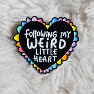 Peut inclure: Épinglette en émail en forme de cœur noir avec le texte "Following My Weird Little Heart" en blanc. L'épinglette a une bordure festonnée colorée avec des nuances de rose, jaune, bleu et vert.