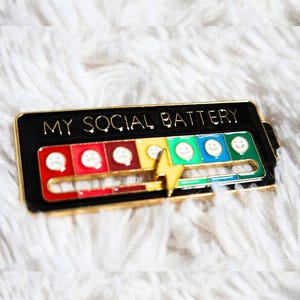 Peut inclure: Une épingle en émail noir avec le texte "MY SOCIAL BATTERY" et un indicateur de niveau de batterie. L'indicateur affiche une gamme allant des visages tristes rouges aux visages heureux verts, avec un éclair jaune au milieu. L'épingle est sur une surface blanche et moelleuse.