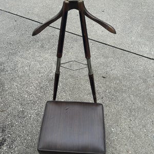Könnte beinhalten: Ein Vintage-Herrendiener mit einem dunkelbraunen Holzrahmen, einem gepolsterten Sitz und einem oberen Bereich zum Aufhängen von Kleidung. Der Ständer hat einen Kleiderbügel und ein kleines Tablett. Die Metallakzente verleihen dem Design einen Hauch von Eleganz.