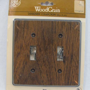 Puede incluir: Una placa de pared de madera de grano marrón con dos aberturas para interruptores y cuatro agujeros para tornillos. La placa está etiquetada como "WoodGrain Designer Collection Wall Plate" y "Maple PWG428D".
