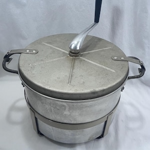Könnte beinhalten: Ein runder, vintage Popcorn-Maker aus Metall mit Deckel und Handkurbel. Der Popcorn-Maker hat eine silberne Oberfläche mit einem schwarzen Griff an der Kurbel. Er verfügt über zwei Griffe an den Seiten und ein Metallband in der Mitte.
