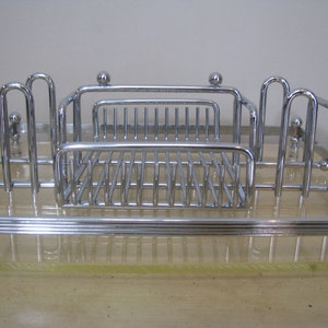 May include: Chrome-plated metal kitchen utensil holder with a clear acrylic base. The holder has a rectangular shape with a raised grid for holding utensils and two vertical dividers.