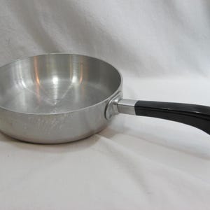 Può includere: Una padella color argento con manico nero. La padella è in metallo e ha una forma arrotondata. Il manico è curvo e fissato alla padella con una staffa metallica. Un piccolo anello di metallo si trova alla fine del manico.