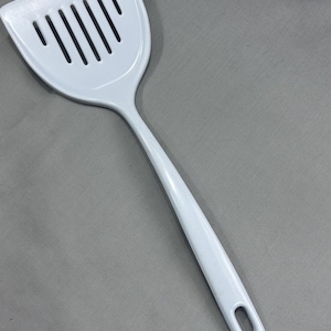 May include: A solid white kitchen spatula featuring a wide, flat head with six vertical drainage slots. The handle is smooth and curved, ending in a small, elongated oval hole designed for hanging the utensil when not in use in the kitchen.