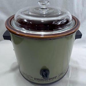 May include: An olive green slow cooker with a brown rim and a clear glass lid featuring a central knob. The base has a black control dial and the text "CROCK-POT SLOW-COOKER" printed in black, accompanied by small graphic illustrations of kitchen items.