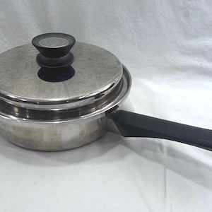 May include: A stainless steel saucepan with a matching lid and a long black handle. The lid features a circular black knob in the centre. The pan has a polished metallic finish and a flat base, designed for stovetop use with a sturdy handle.