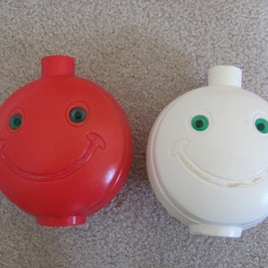 May include: Two plastic piggy banks shaped like smiley faces. One is red with green eyes and the other is white with green eyes.