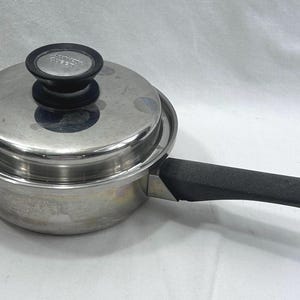 May include: A stainless steel saucepan with a matching lid and a long black handle. The lid features a black knob with a silver centre. The metal surface is reflective and smooth, showing a cylindrical body and a flat, circular lid.