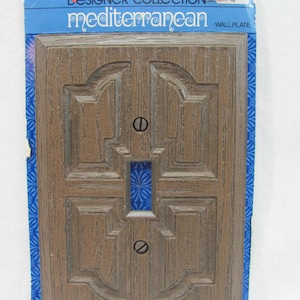 Puede incluir: Una placa de pared de textura de madera marrón con una sola abertura para un interruptor de luz. La placa está empaquetada en un paquete de cartón azul y blanco con el texto "Designer Collection Mediterranean Wall Plate" y el logotipo de General Electric.