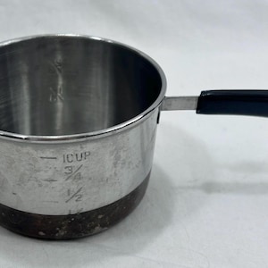 Könnte beinhalten: Ein Messbecher aus Edelstahl mit einem schwarzen Kunststoffgriff und einer Metallöse zum Aufhängen. Die Außenseite weist eingravierte Markierungen für 1 Tasse, 3/4 Tasse, 1/2 Tasse und 1/4 Tasse auf. Der Boden hat eine dunkle, verfärbte Oberfläche.