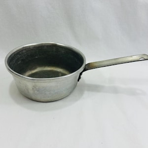 Puede incluir: Una pequeña cacerola vintage de aluminio con un mango largo. La cacerola tiene forma de cuenco redondeado y un solo mango largo que se extiende desde el lateral. El metal tiene un aspecto desgastado y ligeramente deslustrado, lo que sugiere edad y uso. El mango tiene un agujero para colgar.