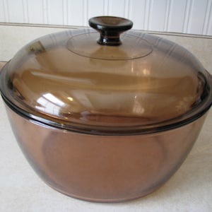 May include: A round, transparent amber-coloured glass cooking pot with integrated side handles and a matching domed glass lid featuring a central knob handle. The cookware has a smooth, polished finish and a wide, deep basin design.