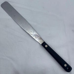 Può includere: Una lunga spatola in metallo dritta con una lama piatta e flessibile in acciaio inossidabile e un manico in plastica nera. Il manico è fissato alla lama con due rivetti metallici color argento e presenta un piccolo foro circolare all'estremità.