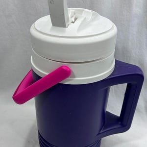 May include: A purple plastic water jug featuring a white screw-top lid with an integrated white straw and a bright pink folding handle. The jug has a sturdy side handle and a ribbed base, designed for carrying liquids with a secure, leak-resistant closure.