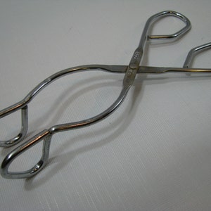 May include: A pair of shiny, silver-colored metal tongs. The tongs have a scissor-like design with looped handles and curved arms that meet at a pivot point. The tongs are likely used for serving food.