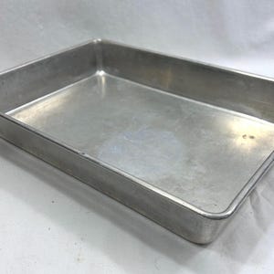 May include: A rectangular metal baking tin with a smooth, silver-toned finish. The tin features straight, vertical sides and a flat base, constructed from a single piece of polished aluminium with rounded corners and a reinforced rim.
