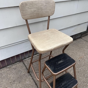 May include: Vintage step stool chair with a light wood seat and backrest, and two black steps. The frame is a bronze color. The chair is designed to provide both seating and a step-up function.