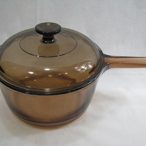 May include: A translucent amber-colored glass saucepan with a matching glass lid. The cookware features a long, flat handle on one side and a small tab handle on the opposite side. The lid includes a circular knob handle in the center.