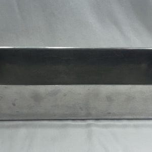 May include: A rectangular, silver metal baking tin with a slightly textured surface. The tin has a handle on each end. The interior is empty, and the tin sits on a white surface.