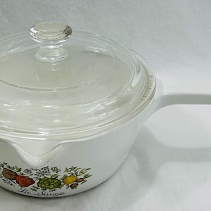 May include: White saucepan with a clear glass lid and a long handle. The pan features a colorful floral and vegetable design with the text "La Sauge" on the side. The lid has a clear knob.
