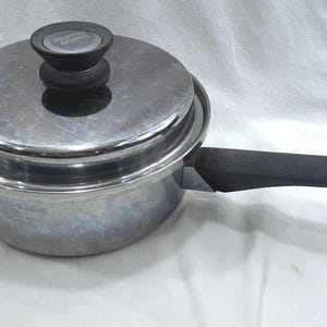 May include: A stainless steel saucepan with a matching lid and a black handle. The lid features a black knob with a circular silver centre. The metal surface is reflective and smooth, showing a cylindrical body and a long, sturdy black handle.