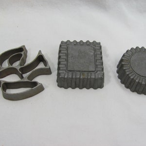 May include: Three vintage metal cake molds. The molds are in the shape of a dove, a square, and a circle. The molds are made of a dark gray metal and have a textured surface.