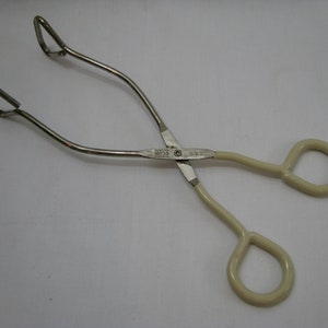 Può includere: Un paio di pinze in metallo con manici color crema e impugnature ad anello. Le pinze sono in metallo color argento e hanno un design a forbice. I manici sono curvi e terminano con un anello.