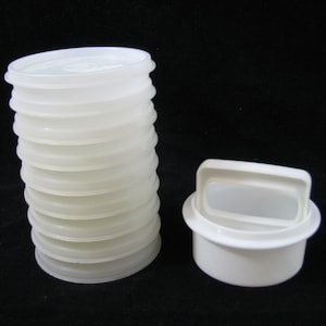 May include: A stack of ten translucent white circular plastic lids and one solid white circular plastic container with a rectangular handle on top. The items are arranged side by side, featuring a smooth finish and simple, functional design.