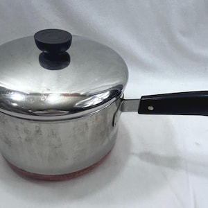 Può includere: Un pentolino in acciaio inossidabile con coperchio abbinato, dotato di un manico in plastica nera e di un pomello nero sul coperchio. Il manico include un piccolo anello metallico all'estremità. La base della pentola ha una finitura color rame.