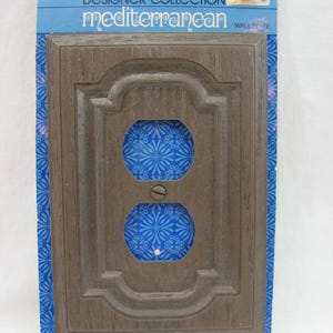 Puede incluir: Una placa de pared de madera marrón con un diseño decorativo. La placa es para una sola toma de corriente y tiene un inserto con estampado azul y blanco. El embalaje dice "Designer Collection Mediterranean Wall Plate" y "General Electric".