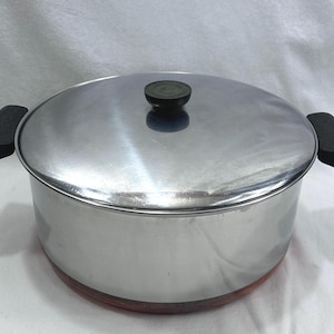 May include: A stainless steel stockpot with a polished silver finish, featuring two black side handles and a matching lid with a central black knob. The base of the pot shows a copper-coloured bottom, providing a contrast to the metallic body.