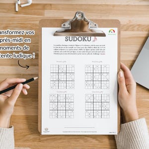 May include: A clipboard holding a Sudoku puzzle sheet with four distinct grids. The text at the top reads SUDOKU, followed by instructions in French. A black pencil is positioned to the left of the clipboard, which is placed on a wooden surface.